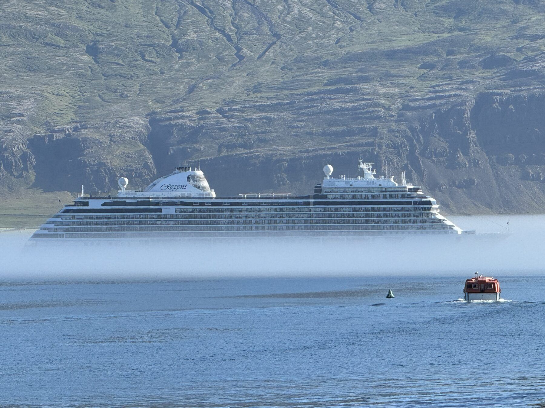 A cruise ship at anchor in the fjord, sea mist across the water and a tender heading ashore