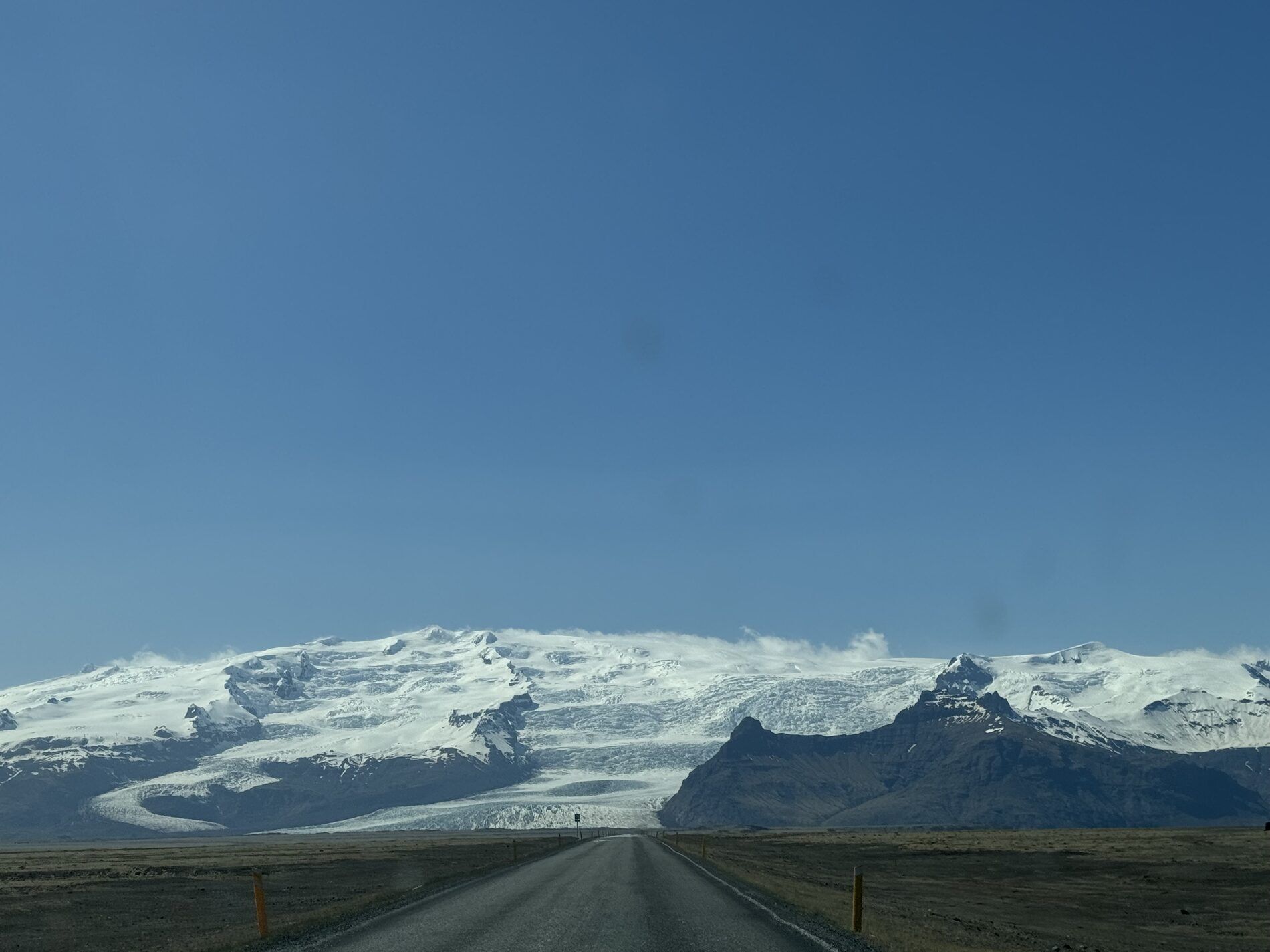 An empty road running straight toward the snow-covered Vatnajökull ice cap, outlet glaciers spilling down between dark peaks