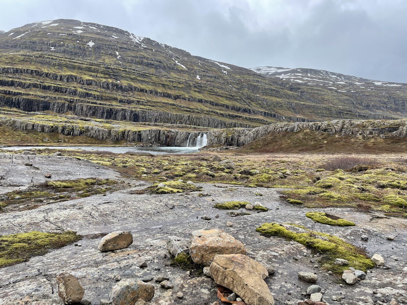 A waterfall dropping over layered basalt into a river below a moss-covered mountainside